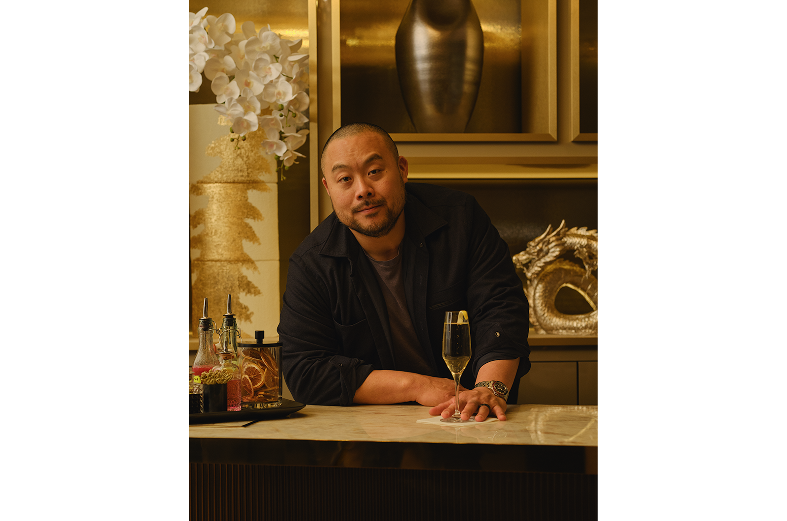 Chef David Chang at a gold-accented bar with a champagne glass, floral decor, and elegant sculpture.