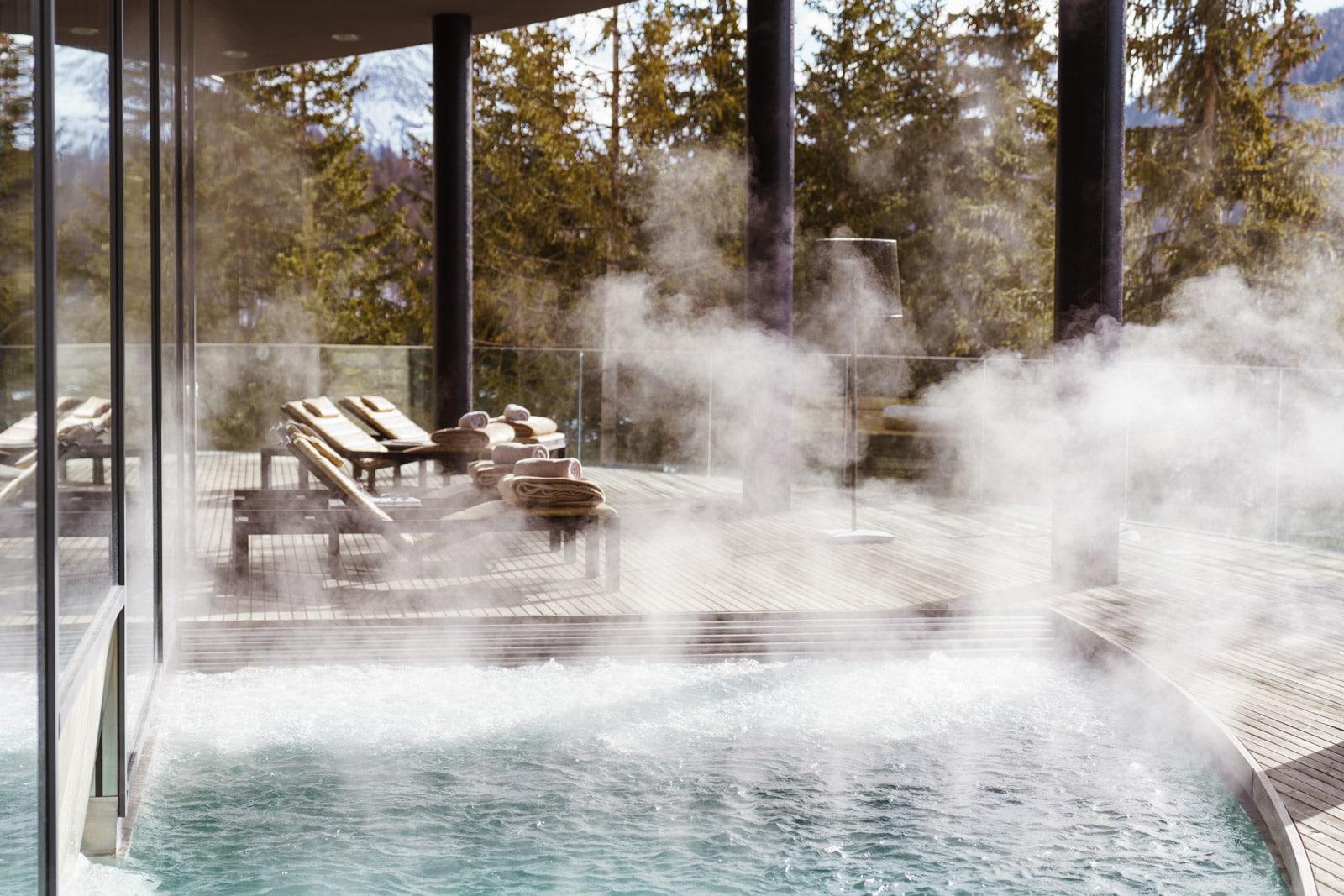 Steam rises above an indoor spa, which has windows looking into a pine forest.