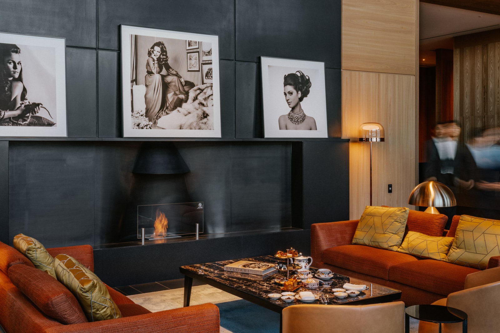 Stylish lounge with orange sofas, modern fireplace, and black-and-white portraits on dark walls.