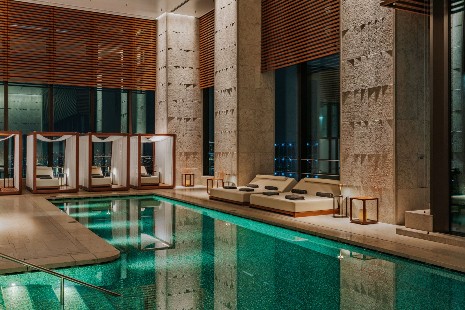 Indoor pool with turquoise water, beige lounge beds, and private cabanas under warm lighting.