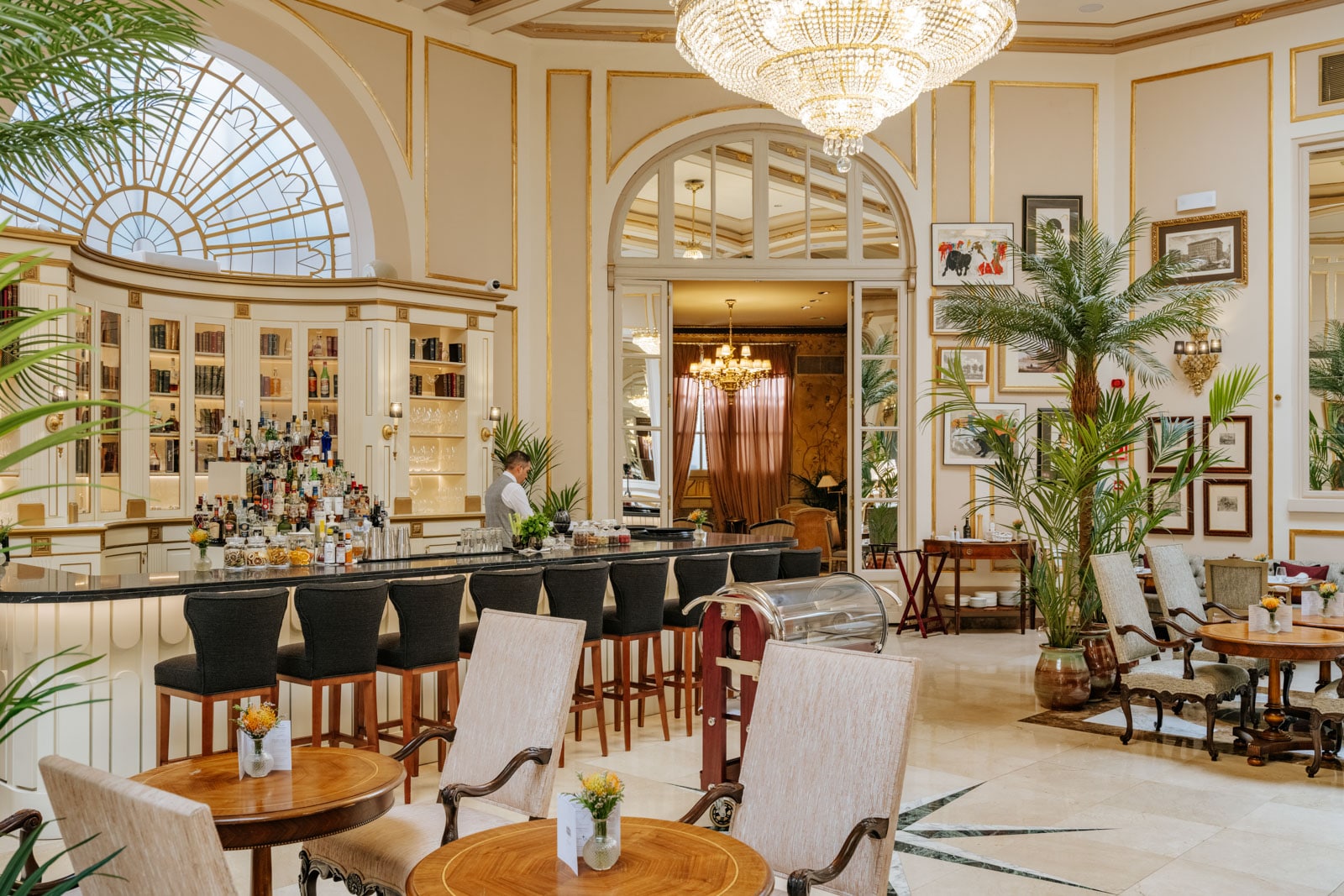Bright bar lounge with arched windows, black chairs, gold trim, green plants, and framed art on cream walls.