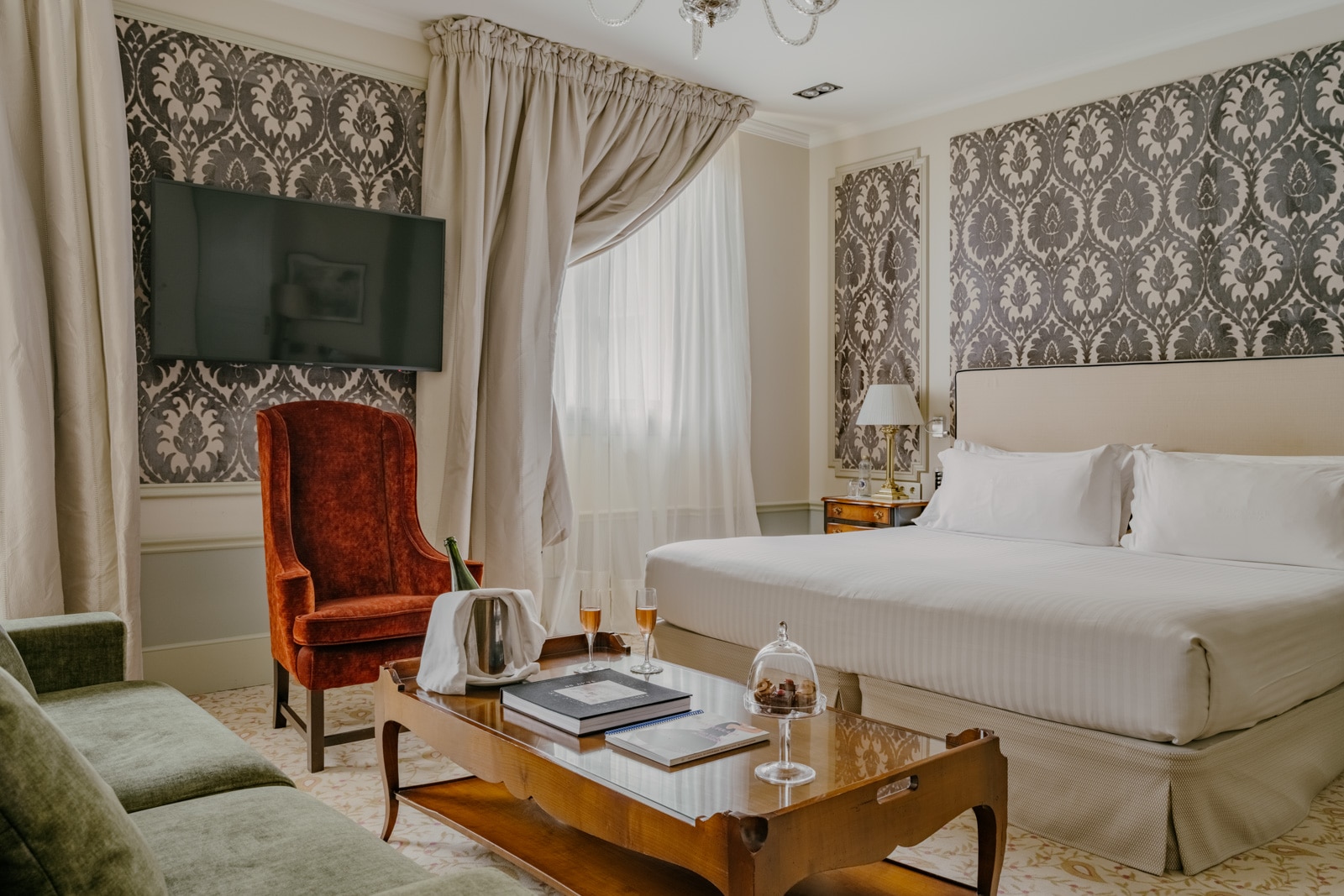 Stylish hotel room with patterned gray walls, white bed, velvet red chair, green sofa, and wood table.