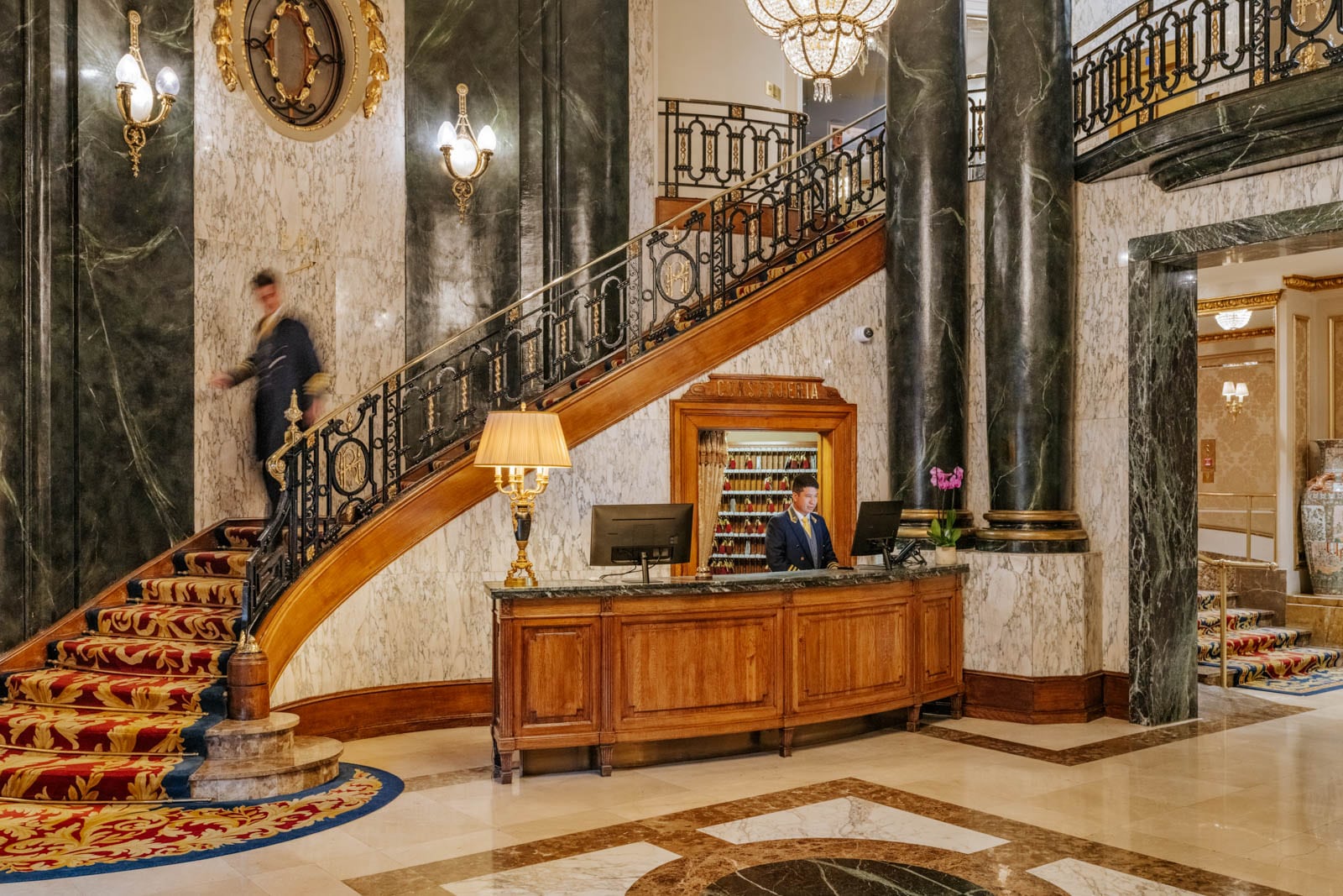 Grand marble lobby with green columns, gold accents, red and gold carpet, and a wood reception desk.