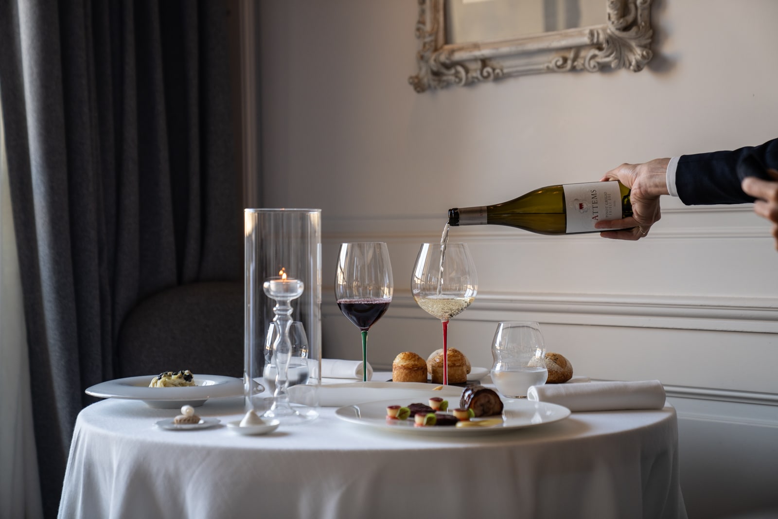 A hand pours white wine into a glass at a table set with gourmet food, wine glasses, bread, and a candle in a restaurant.