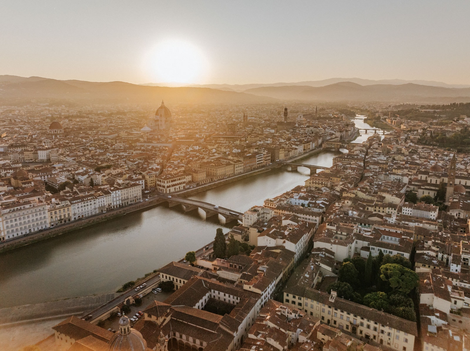 Aerial view of Florence at sunset, showing the Arno River, bridges, and city rooftops with the sun low in the sky.