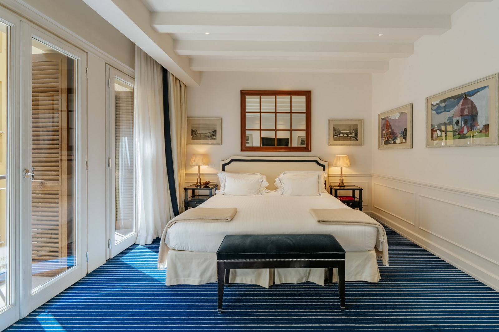 Spacious hotel room with a large bed, striped blue carpet, and artwork on the walls, illuminated by natural light.