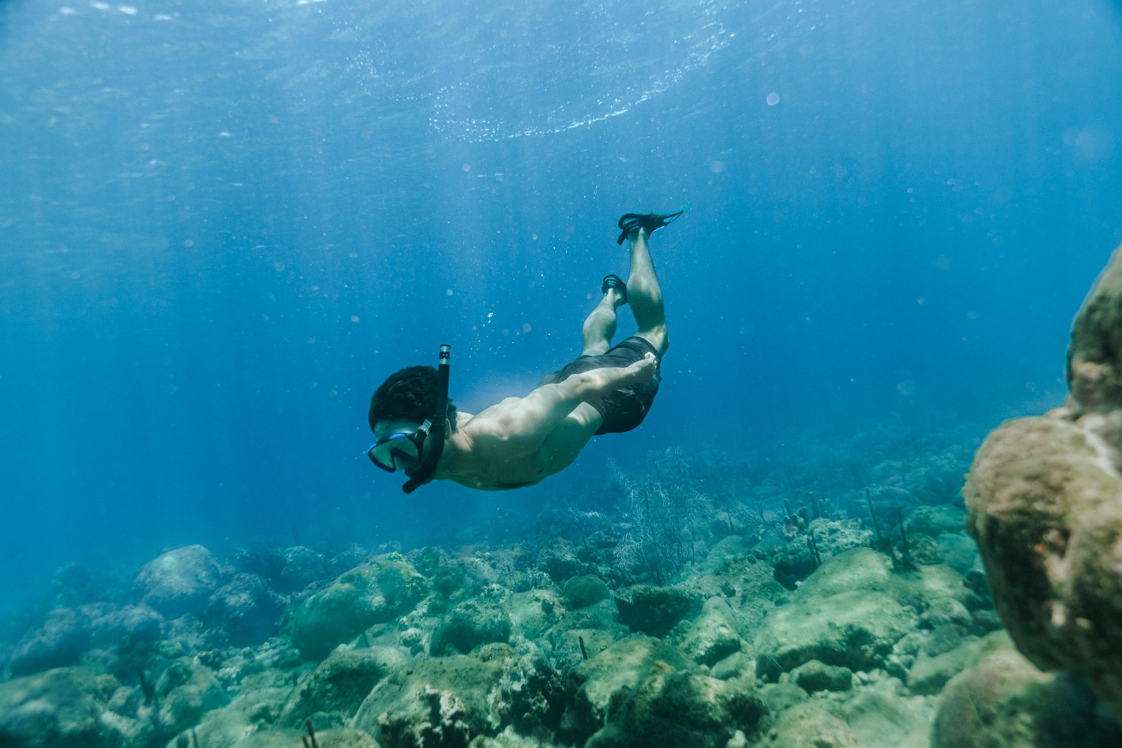 Underwater swimmer with fins over coral rocks in clear blue sea, sunlit bubbles above.