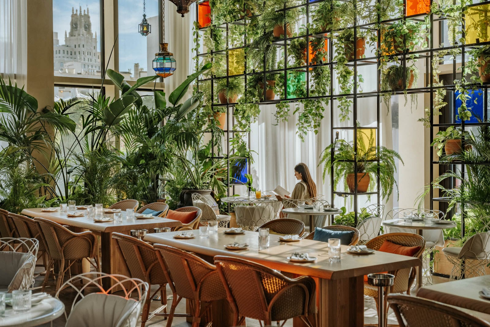 Sunlit restaurant with lush greenery, colorful glass panels, and a woman reading at a table.