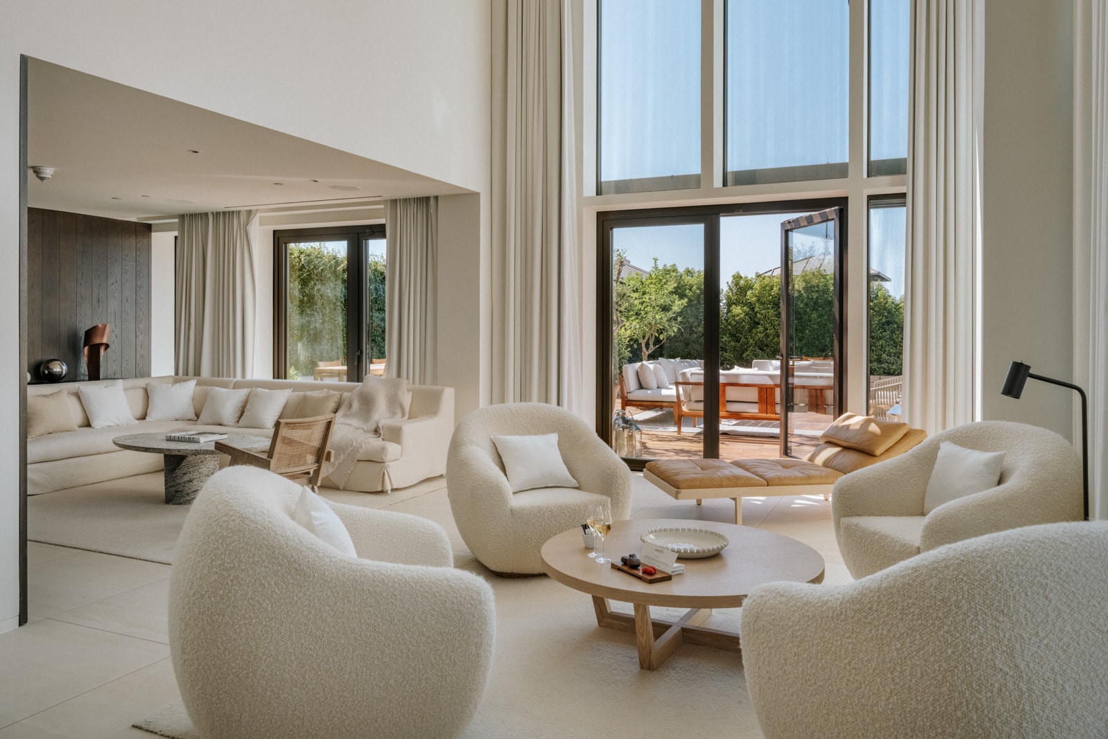 Bright, modern living room with cream armchairs, a round coffee table, large windows, and a view of an outdoor patio.