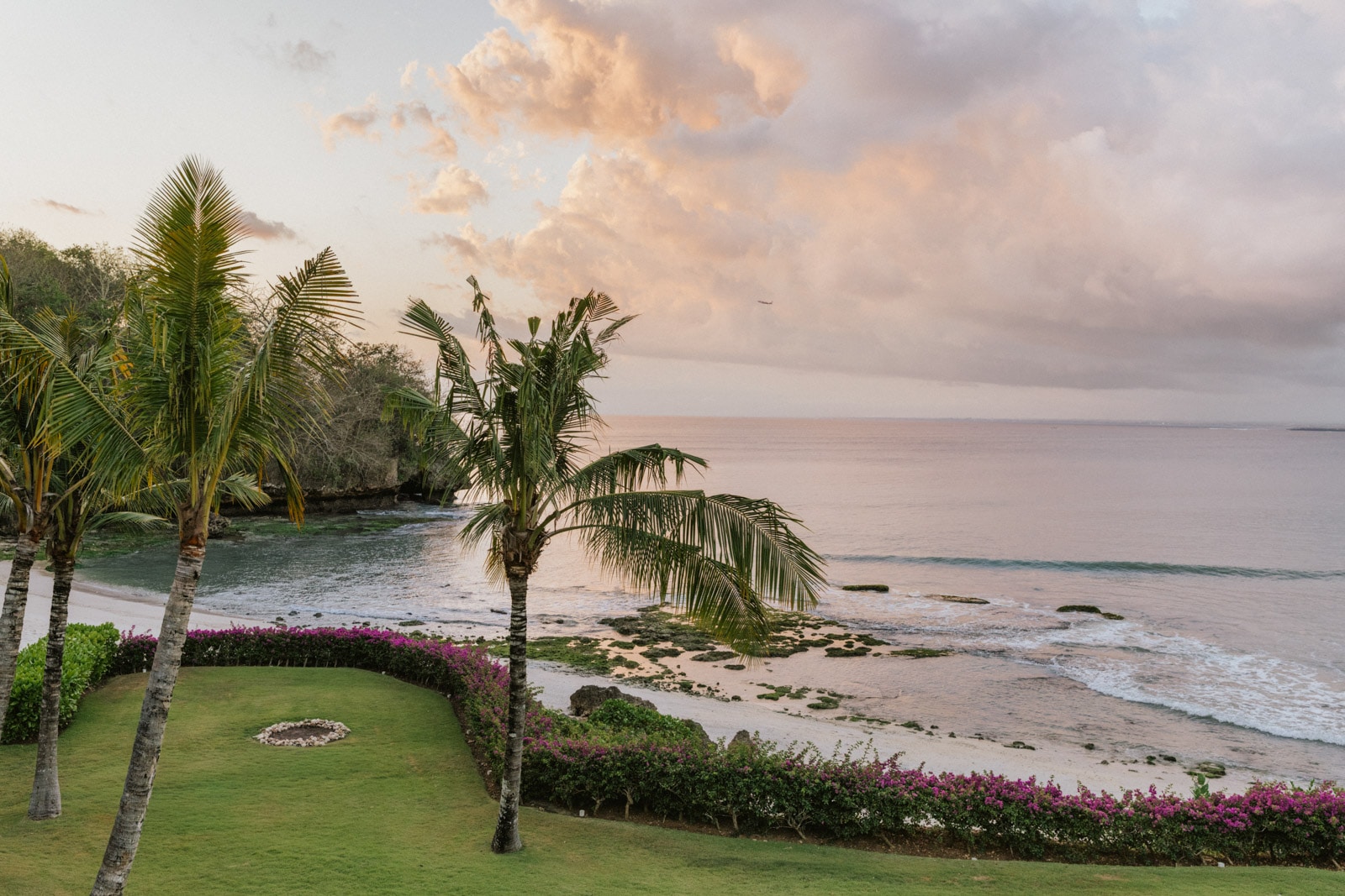 Palm trees and lush lawn overlook a rocky shoreline and calm ocean at sunset.