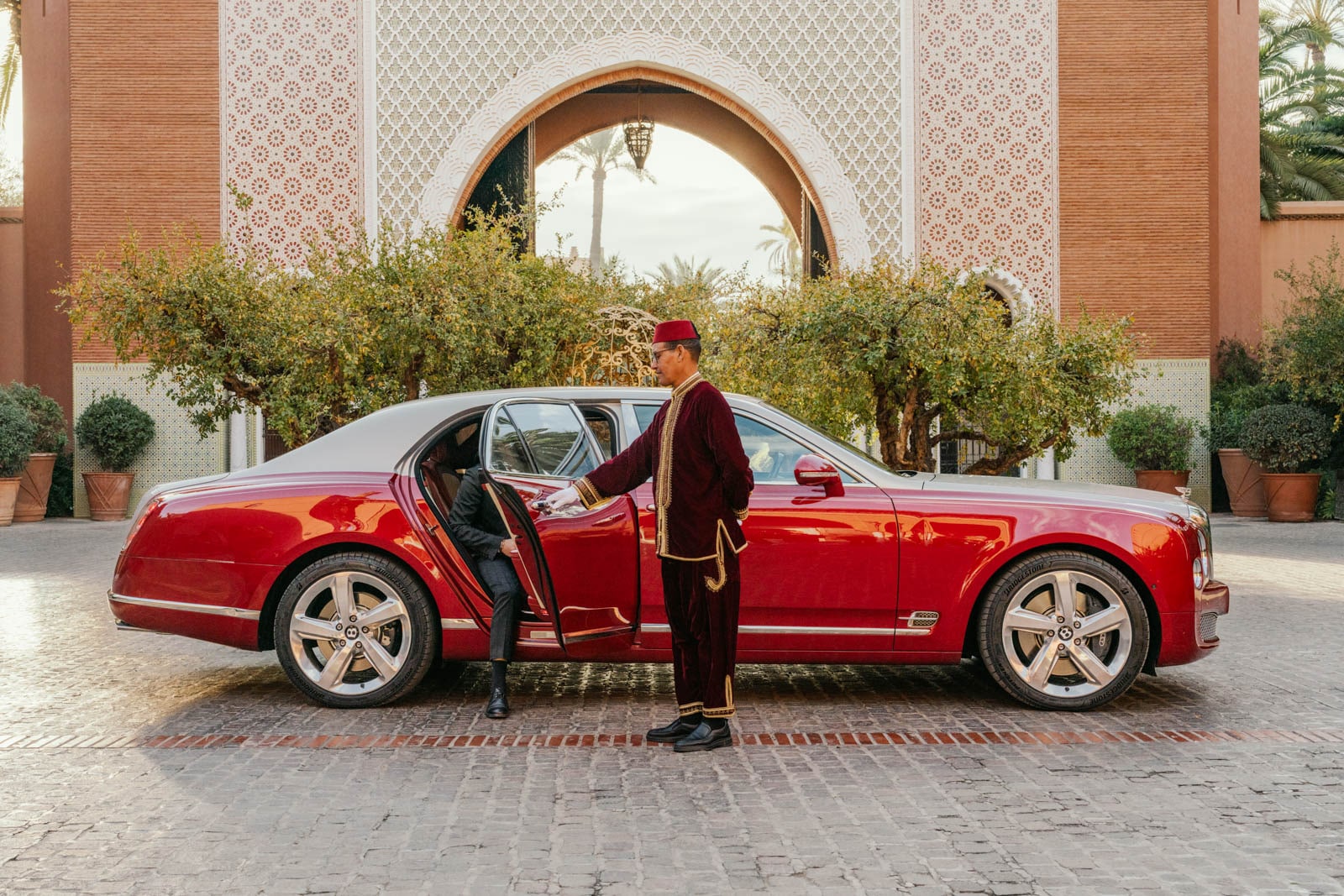 Red luxury car with silver roof parked on cobblestone, doorman in burgundy uniform opens door for guest.