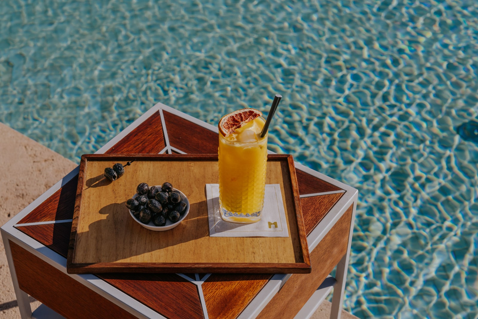 Poolside wooden tray with a yellow citrus drink and a bowl of black olives, beside sparkling blue water.