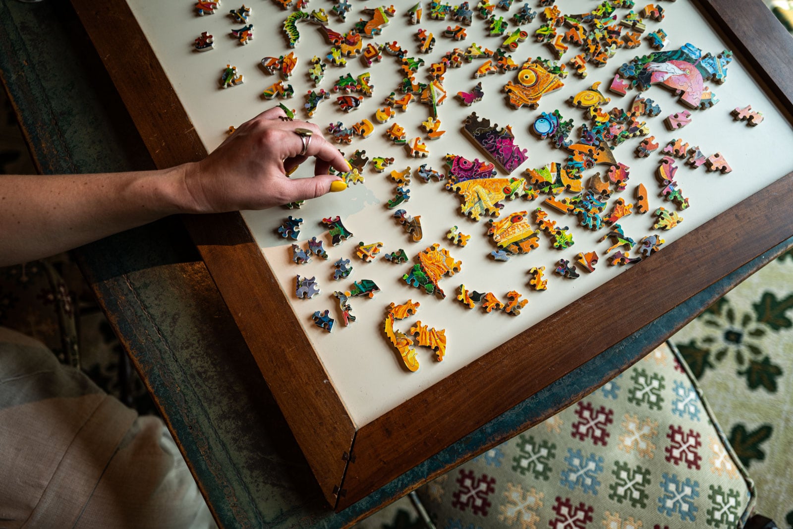 Hand assembling colorful puzzle pieces on a wooden table, patterned chair and green carpet below.