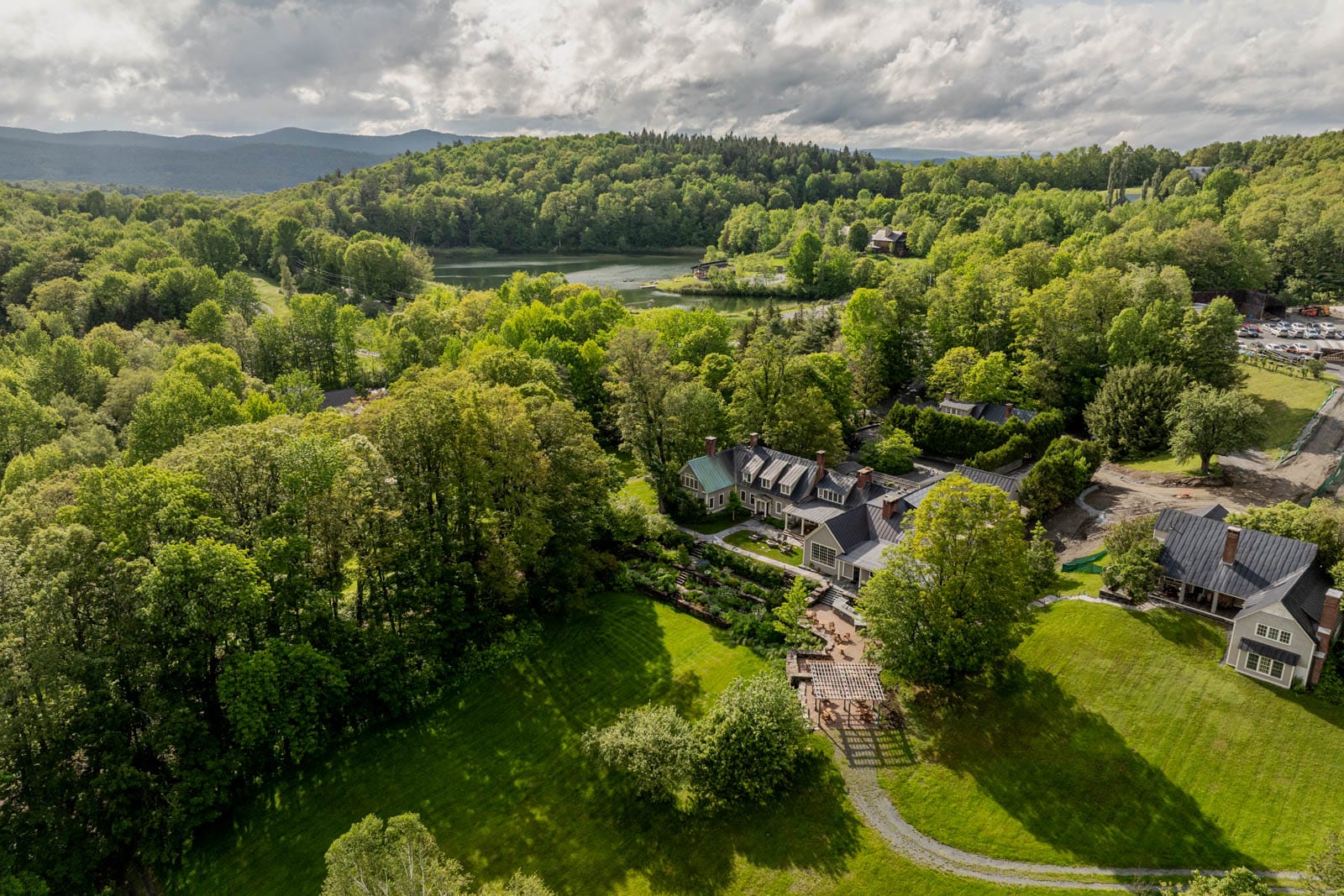 Aerial view of gray farmhouse, gardens, and lake, surrounded by rolling green hills and dense forest.