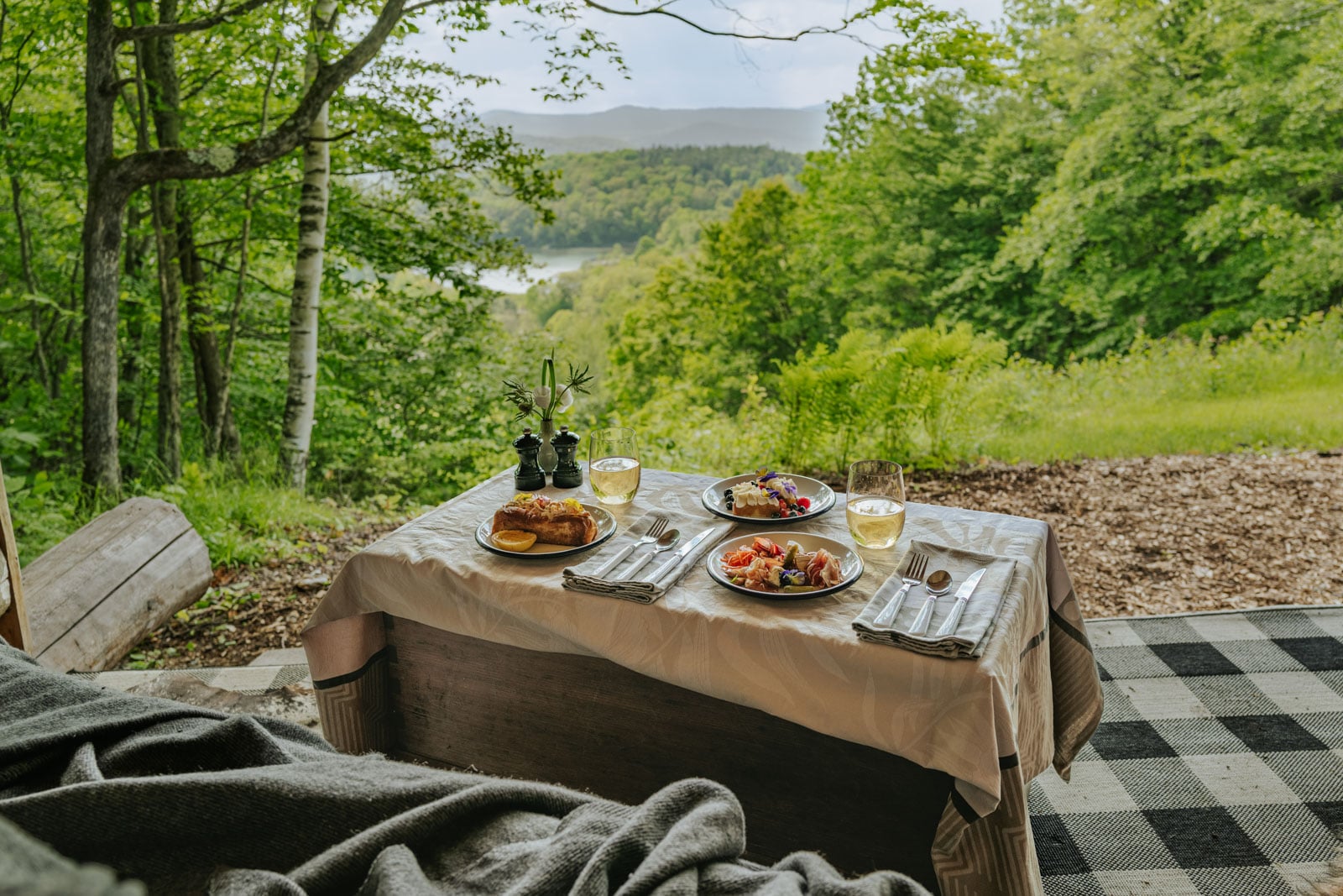 Outdoor breakfast table with pastries, fruit, and wine, overlooking green forest and distant lake.