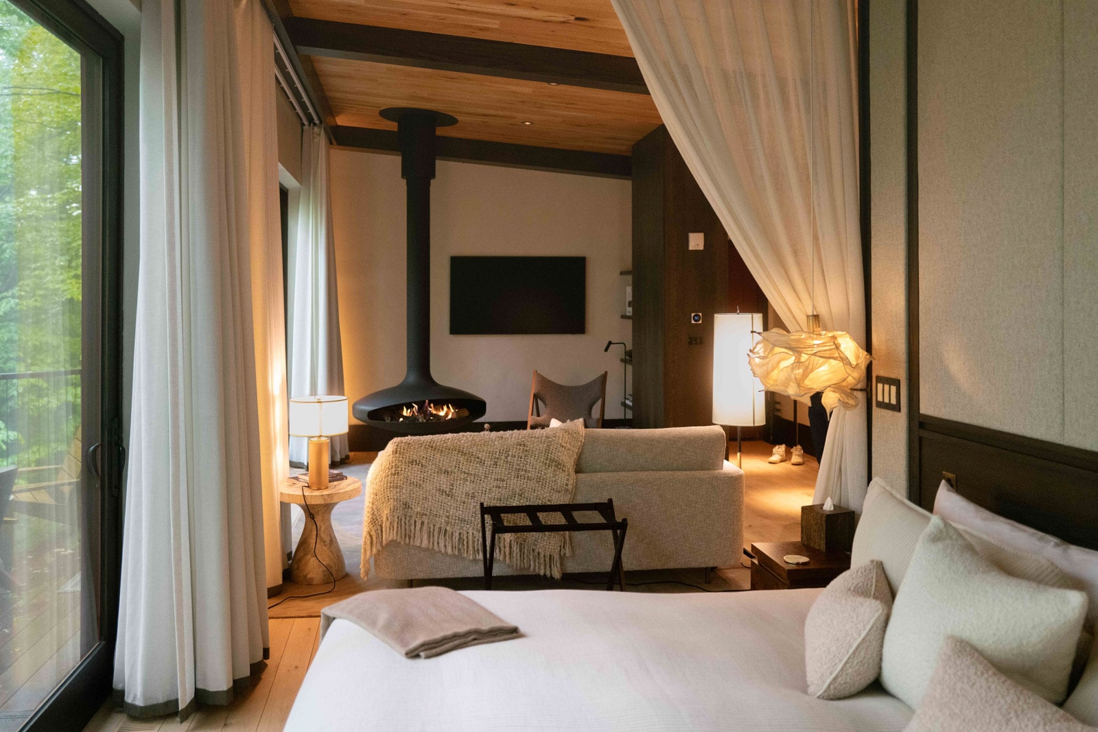 Modern suite with white bedding, soft beige pillows, fireplace, and large windows overlooking green forest.