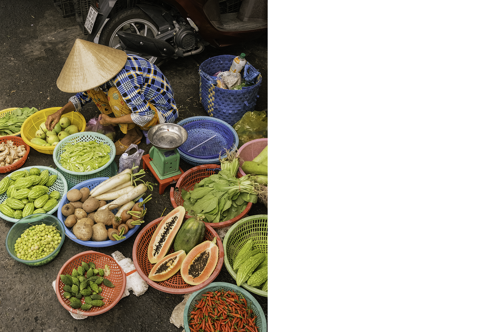 A street vendor in a conical hat sorts produce among bright baskets of fruits and vegetables.