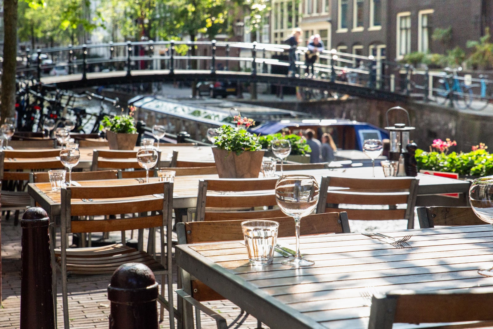 Outdoor café tables set along a canal with a bridge and bicycles in the background.