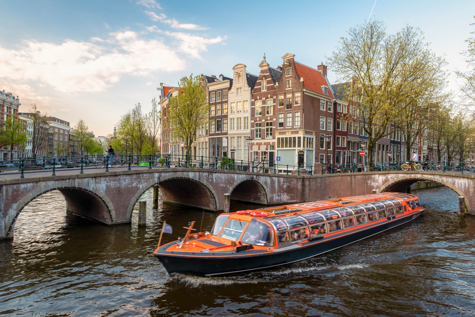 Canal boat cruising under a bridge with traditional Dutch houses and trees lining the water.