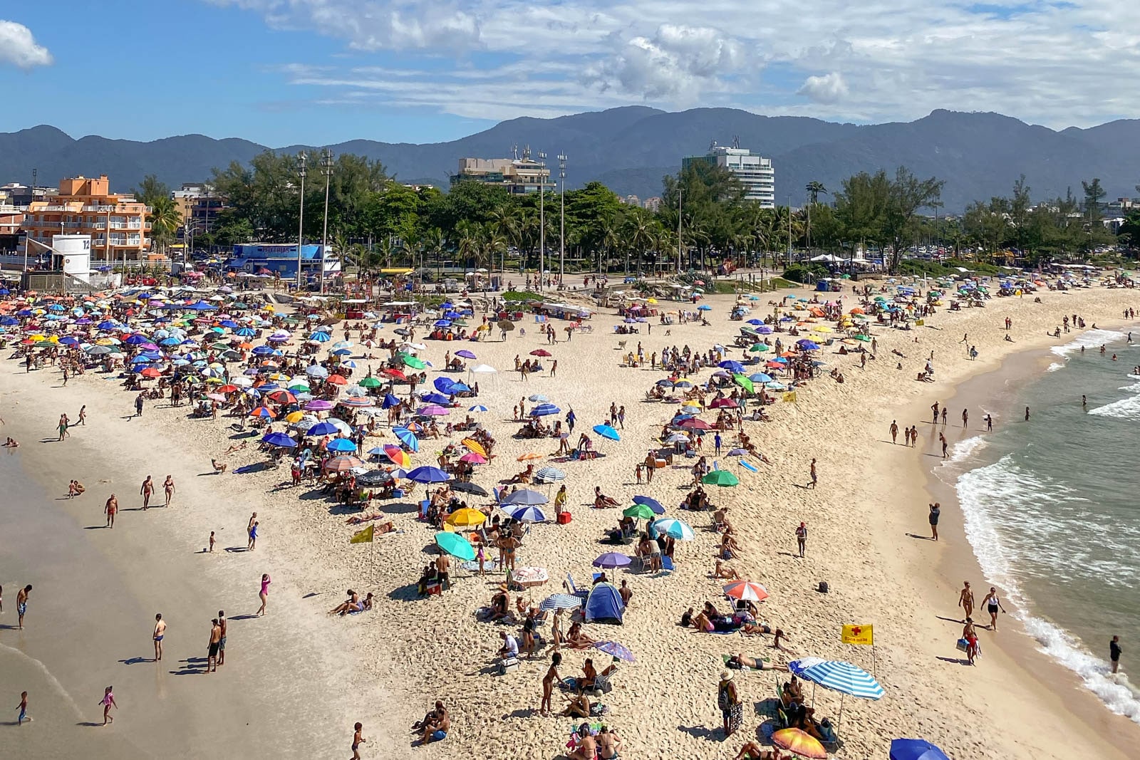 Crowded sandy beach with colorful umbrellas, people sunbathing, and city buildings in the background.
