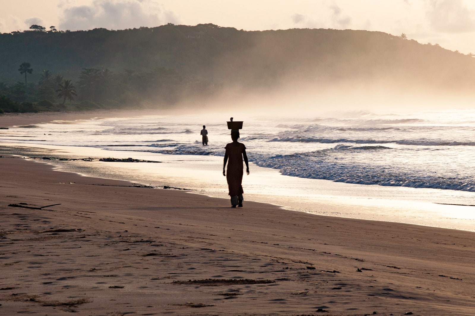 Silhouetted person walking on a misty beach at sunrise, with gentle waves and distant hills.
