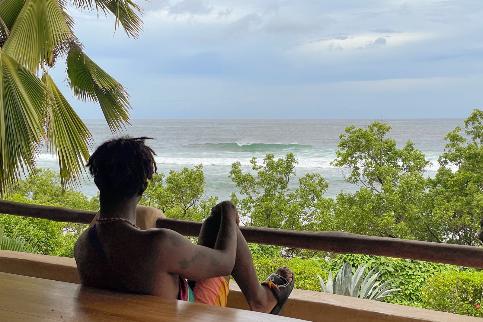 Person relaxing on a wooden deck, looking out at green trees and rolling ocean waves under a cloudy sky.