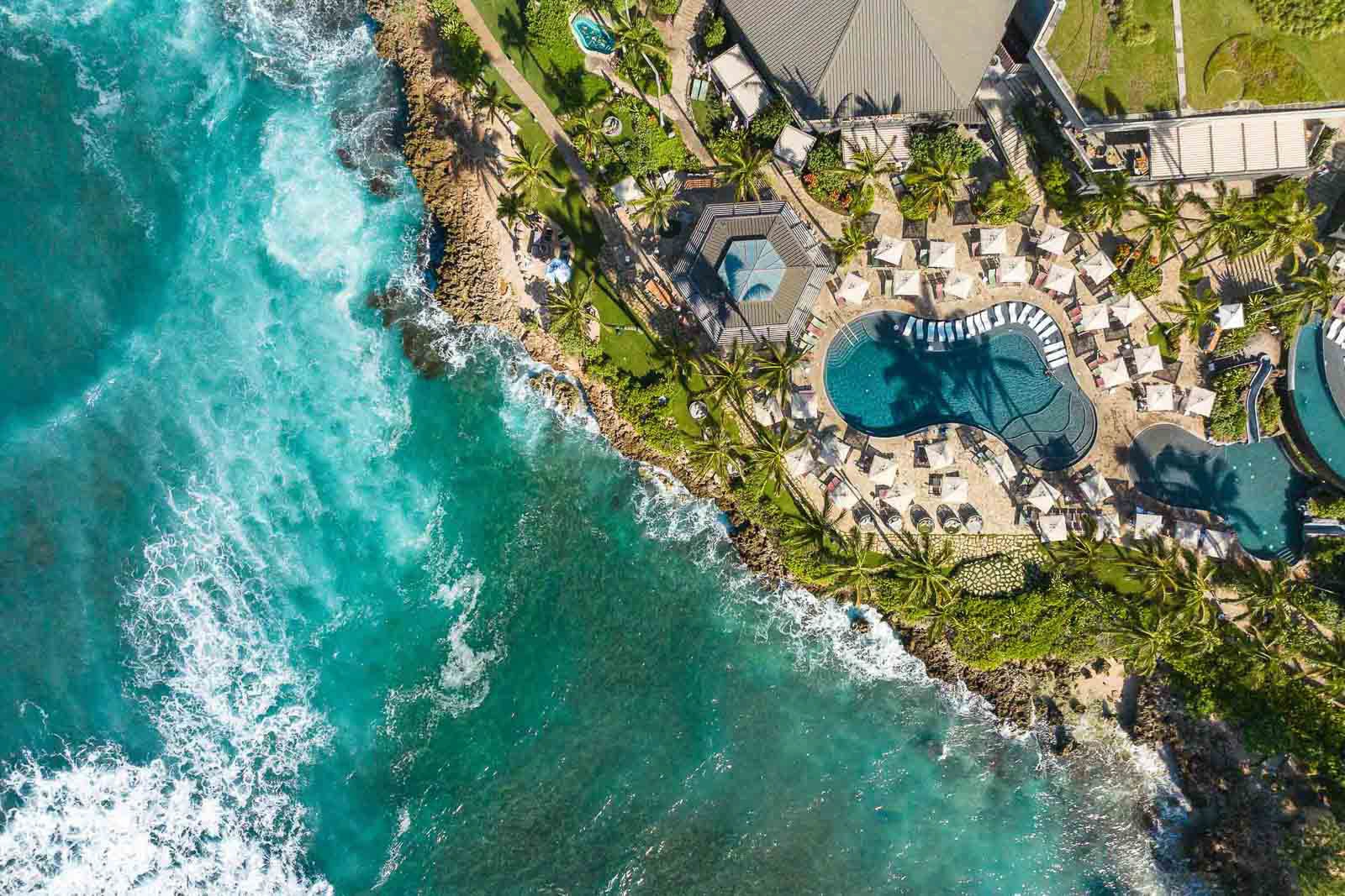Aerial view of turquoise ocean waves crashing against rocky shore, with a pool and palm trees nearby.