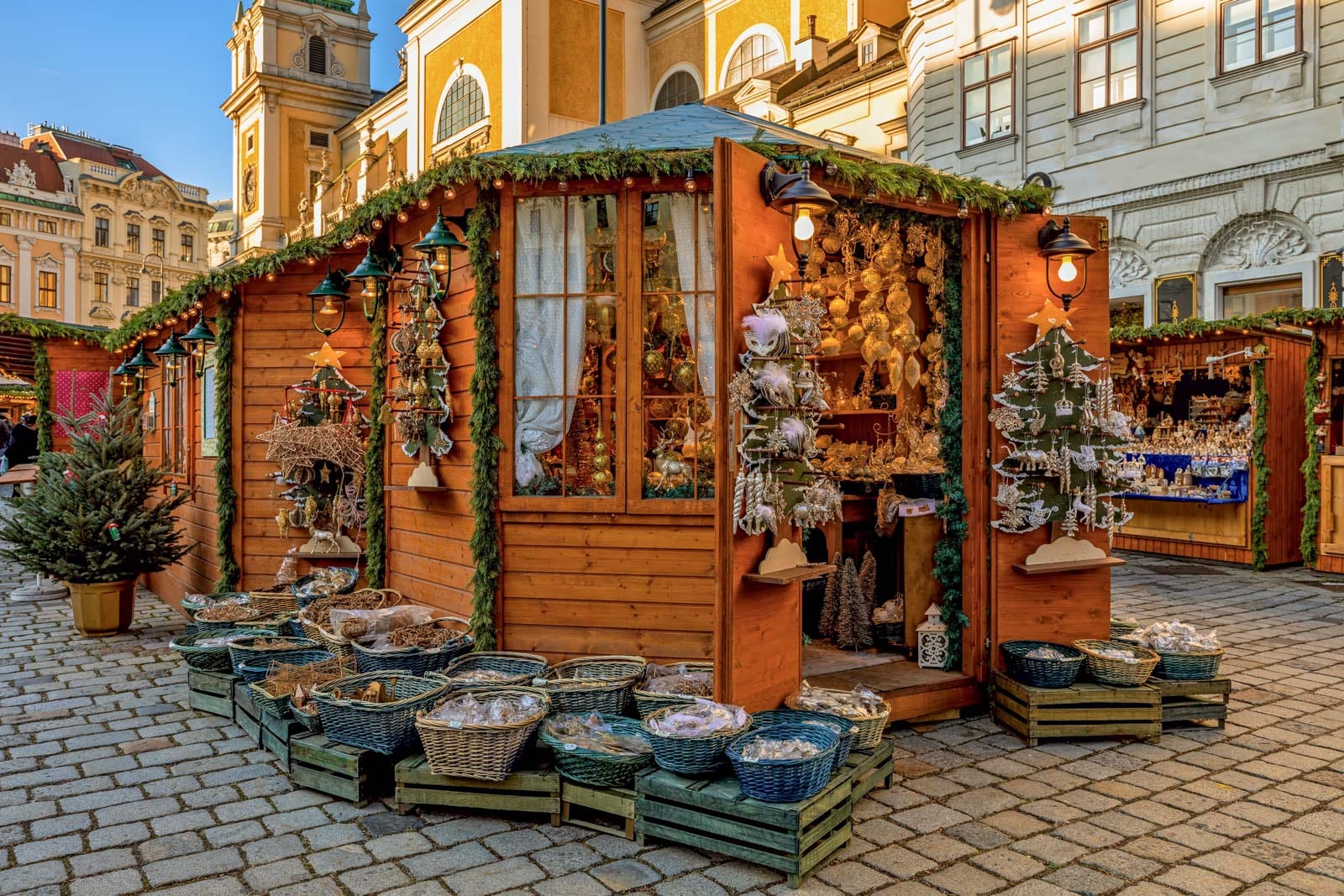 Wooden holiday market stall with green garland, baskets, and ornaments, set on a cobblestone street.