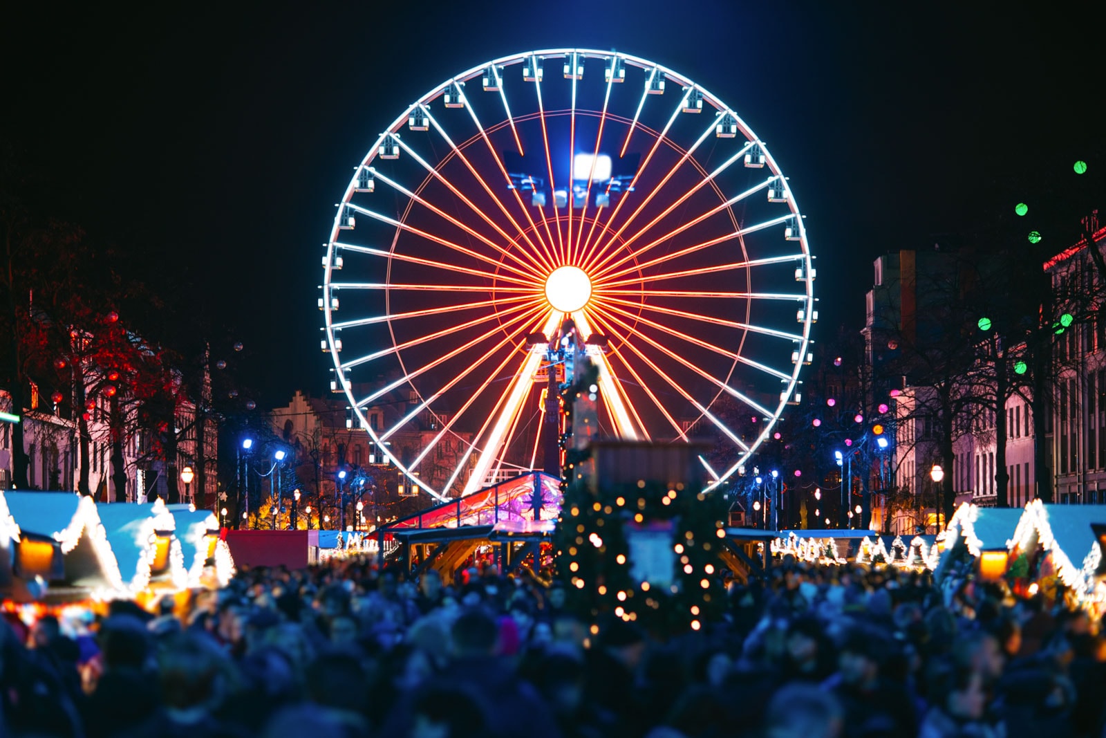 Ferris wheel glowing red and blue at night above a festive market, with crowds and lighted stalls.
