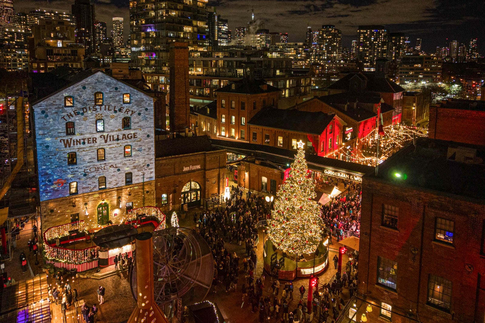 Outdoor winter village at night with a lit Christmas tree, brick buildings, and city lights in the background.