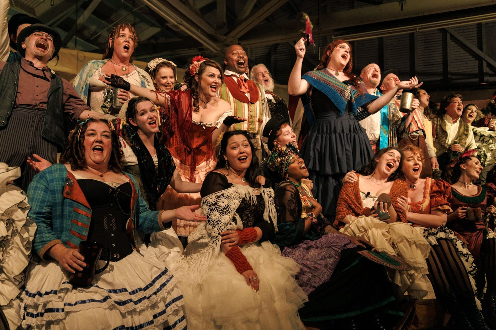 Group of people in colorful period costumes, including dresses with lace, shawls, and striped skirts, gathered indoors.