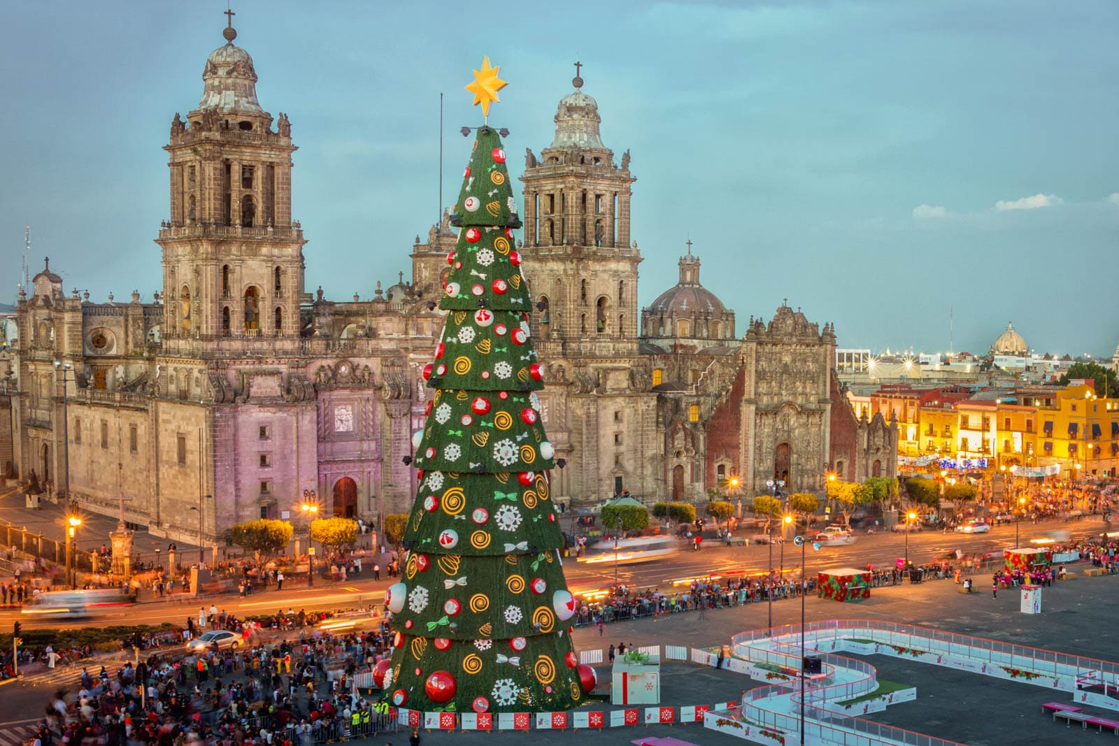 Giant green Christmas tree with ornaments and a yellow star in front of a historic stone cathedral at dusk