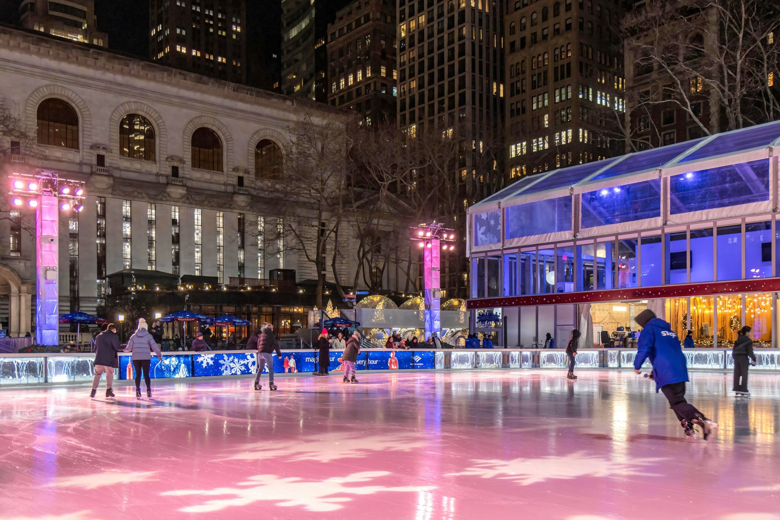 Outdoor ice skating rink with pink and blue lights, skaters, and tall city buildings in the background.