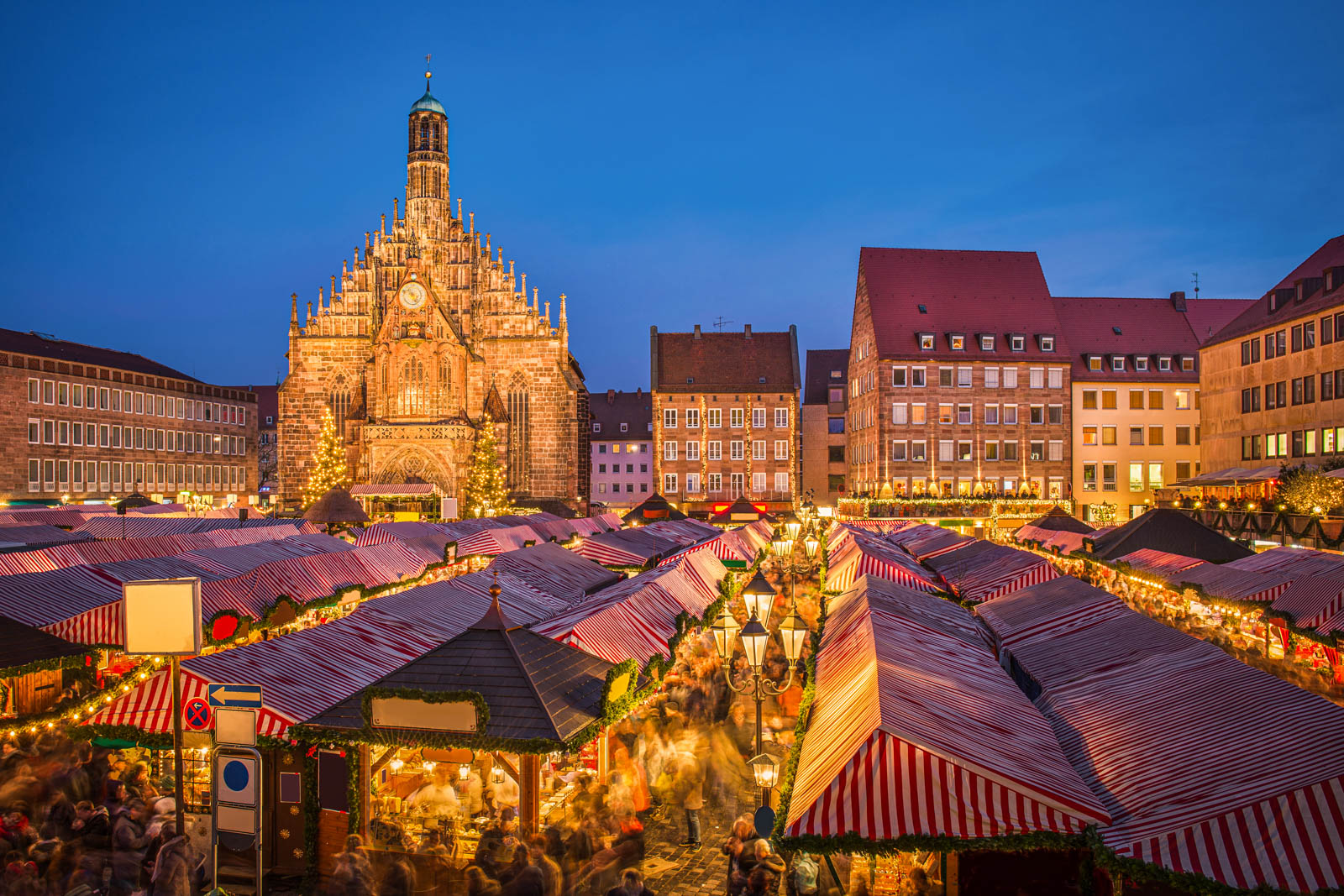 Christmas market with red and white striped roofs, glowing lights, and a Gothic church at dusk.