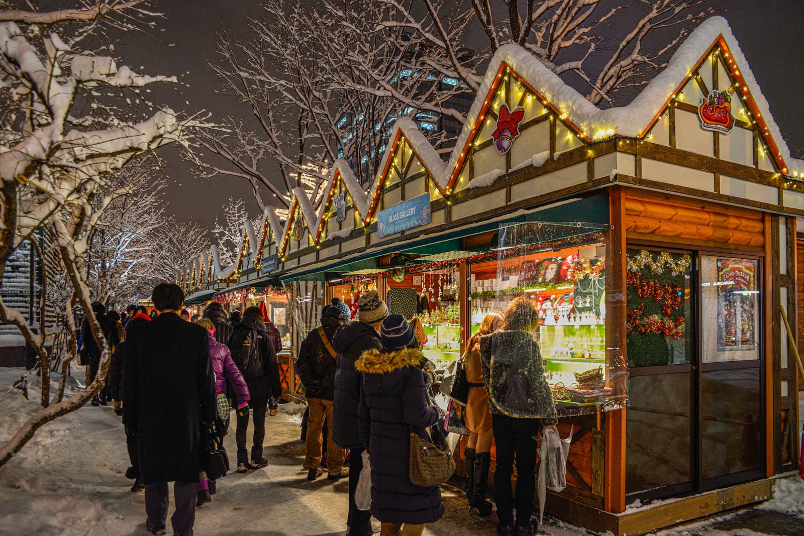 Snowy holiday market with peaked roofs, yellow lights, and people in dark winter coats shopping at night.