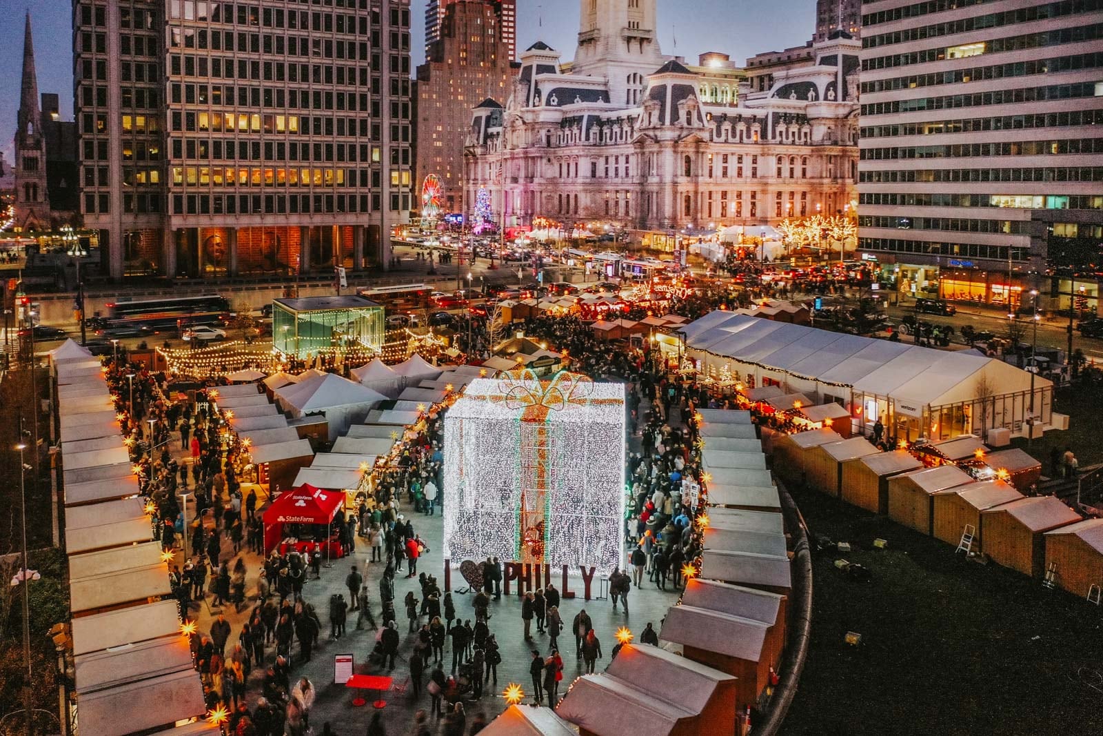 City holiday market with white tents, glowing lights, and a giant illuminated gift box in the center.
