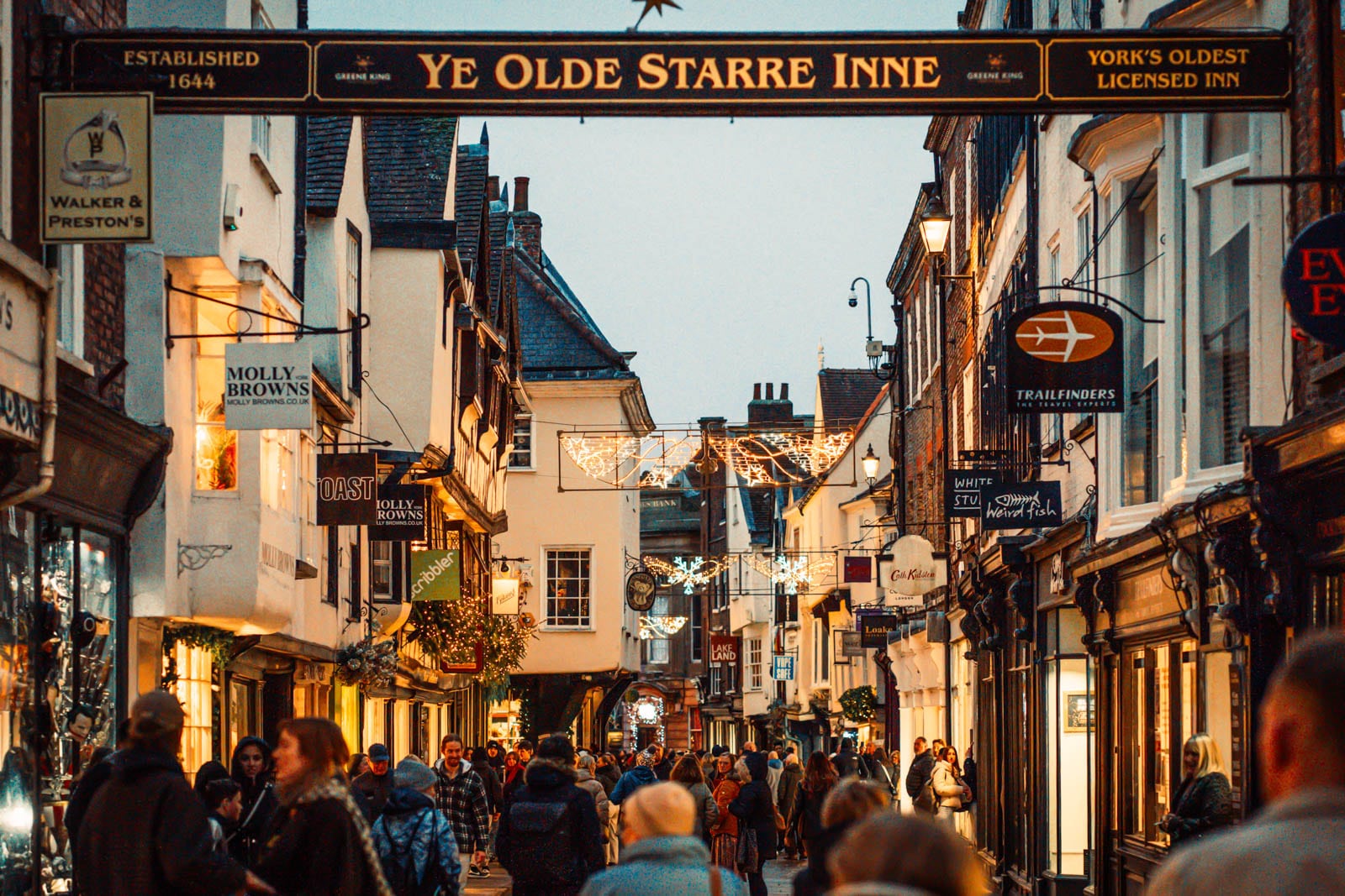 Historic street with festive lights, shop signs, and crowds under a black and gold 