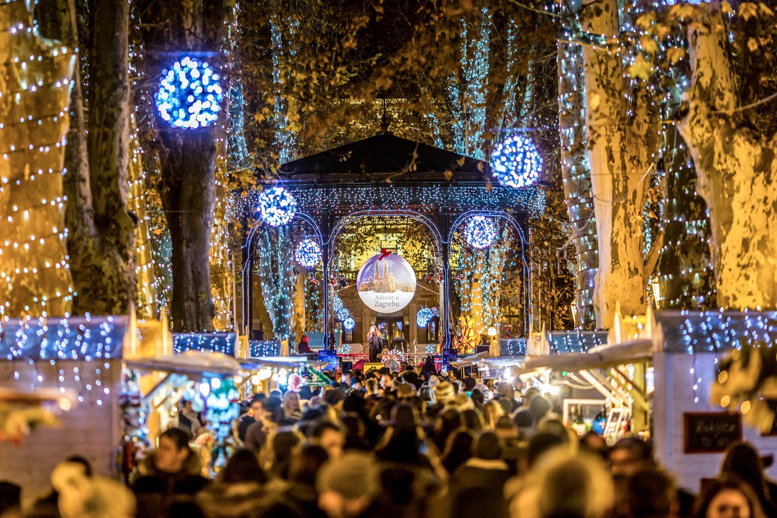 Outdoor market with blue and white holiday lights, glowing trees, and crowds in winter coats at night.