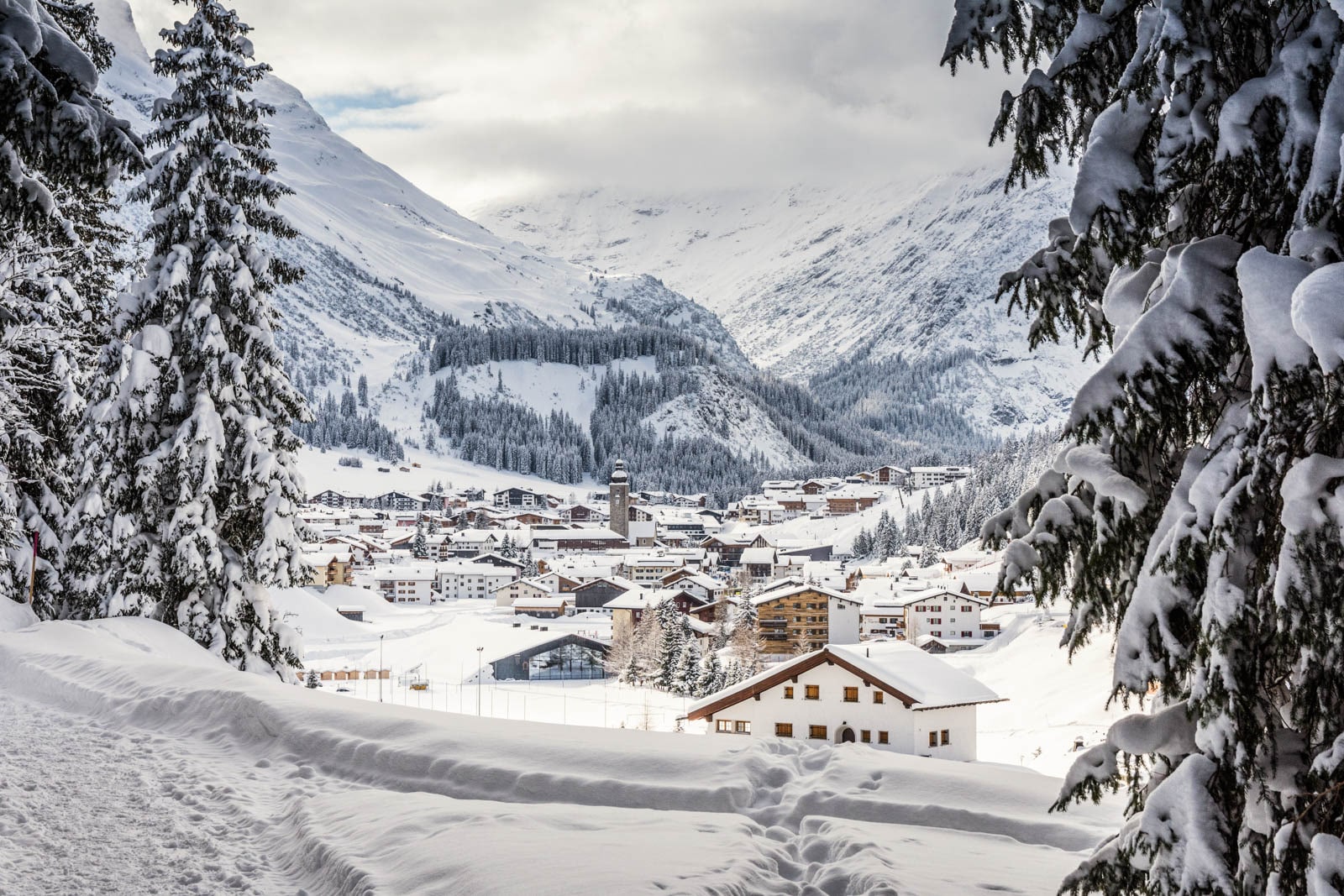 View of Lech, Austria during winter time. Snow-covered alpine village nestled in a valley, surrounded by pine trees.