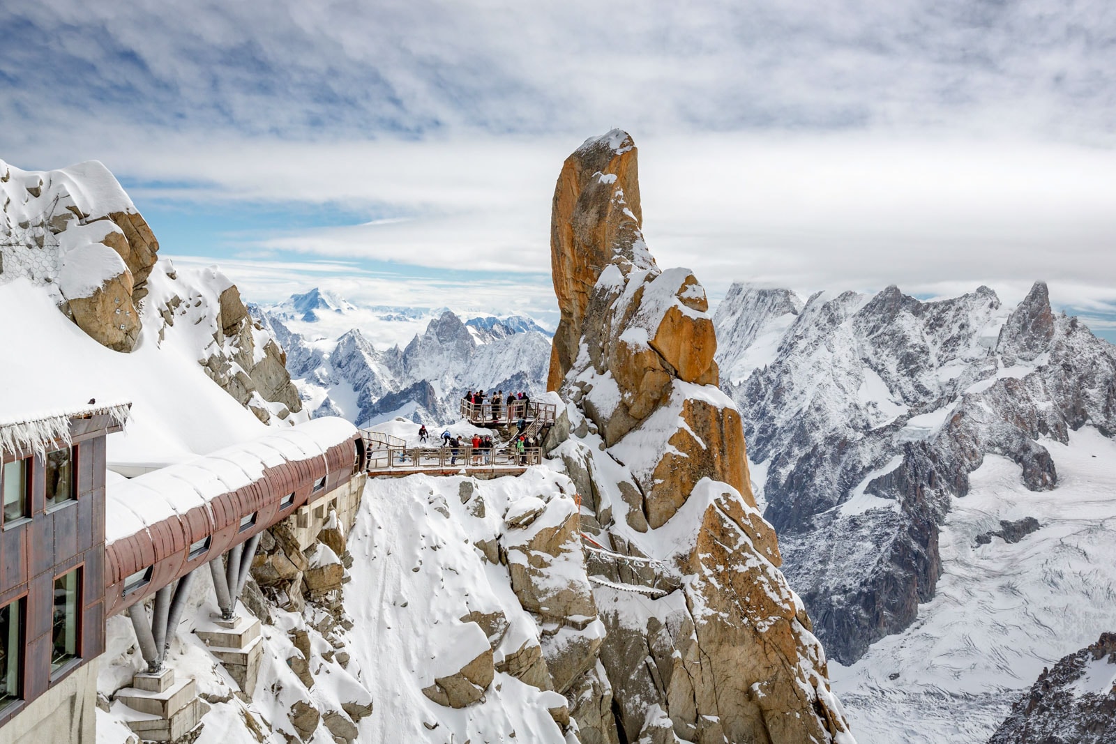 Observation deck on a snowy mountain peak with visitors, rugged cliffs, and panoramic views of surrounding mountains.