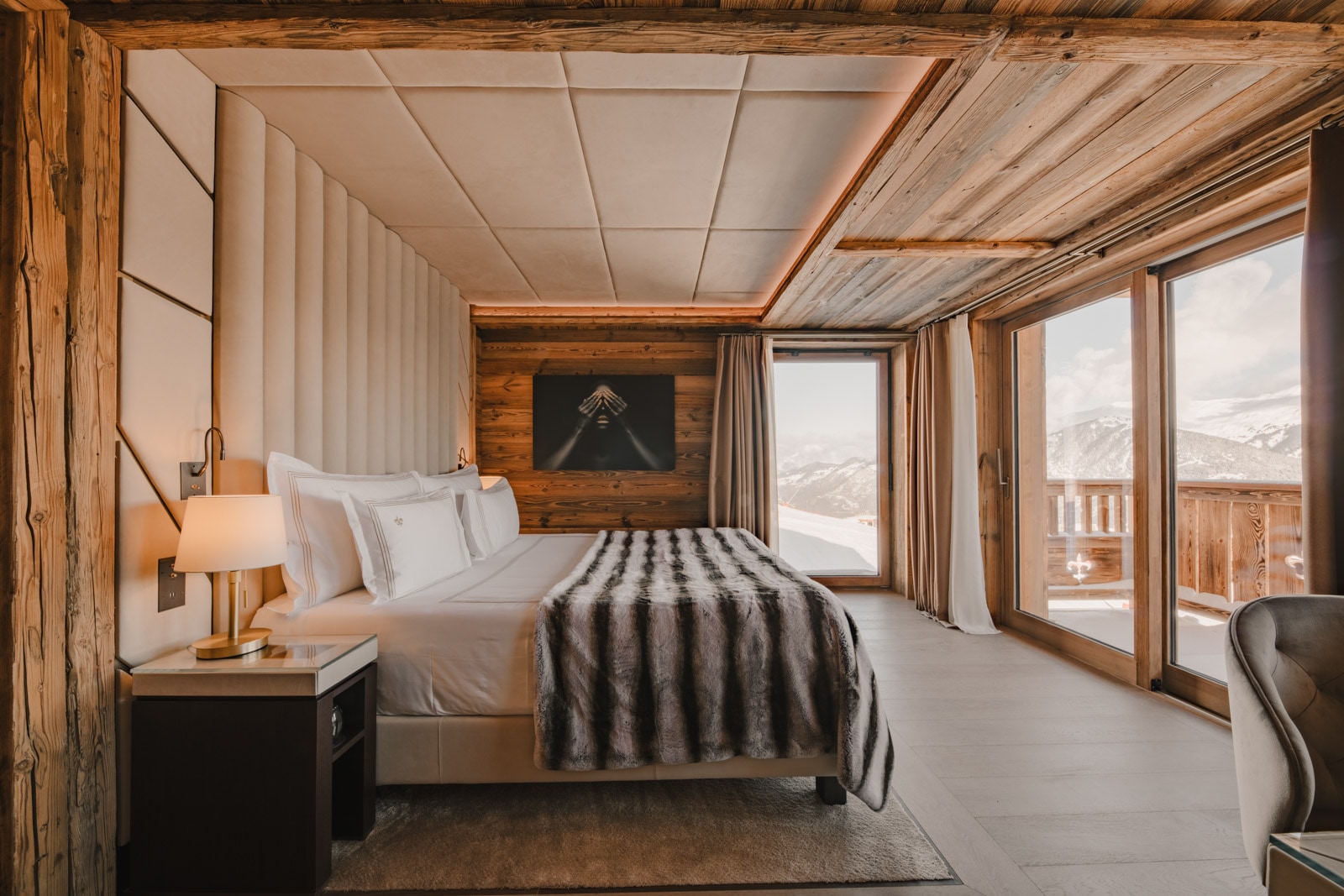 Cozy hotel room with a large bed, wood-paneled walls, and windows overlooking snowy mountains and a balcony.