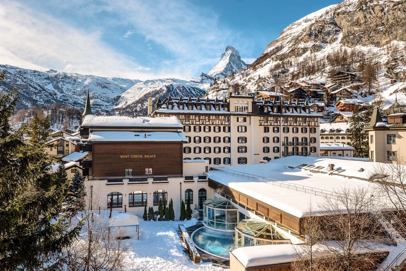 Historic hotel building with snow-covered rooftops and an outdoor pool, set against a backdrop of mountains.