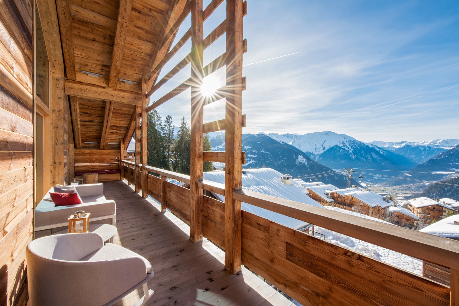 Wooden chalet balcony with modern chairs, sun shining through beams, and scenic views of snowy mountains and ski lifts.