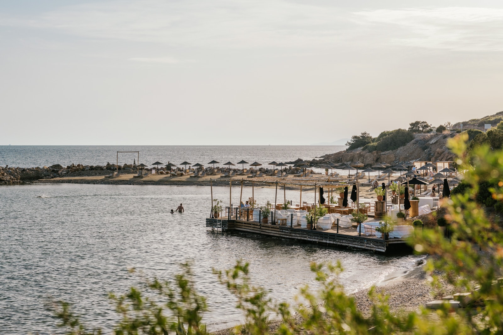 Sunlit seaside cove with black umbrellas and a wooden pier lounge over calm blue water.