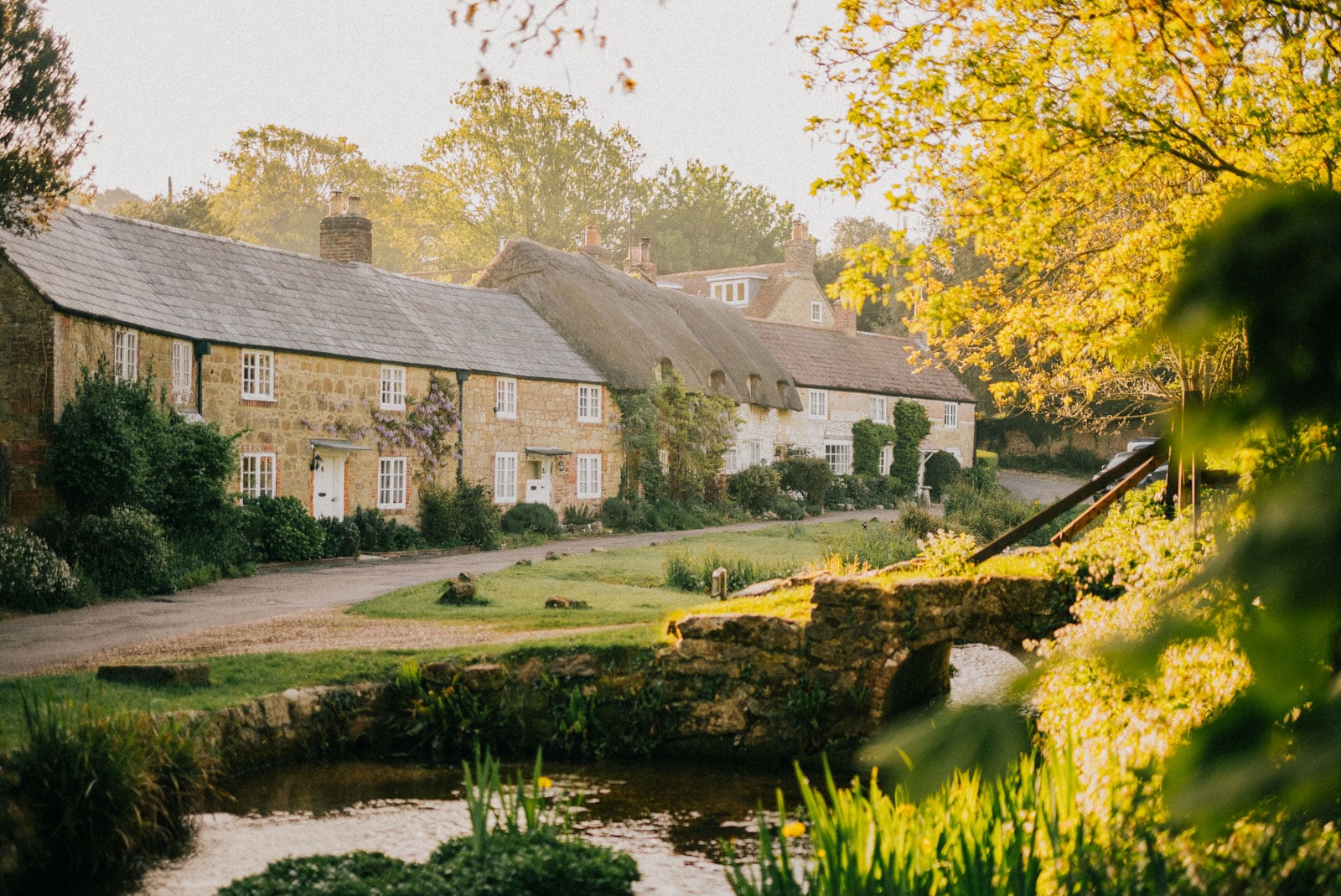 Sunlit village with thatched cottages, stone bridge, and a gentle stream framed by greenery.