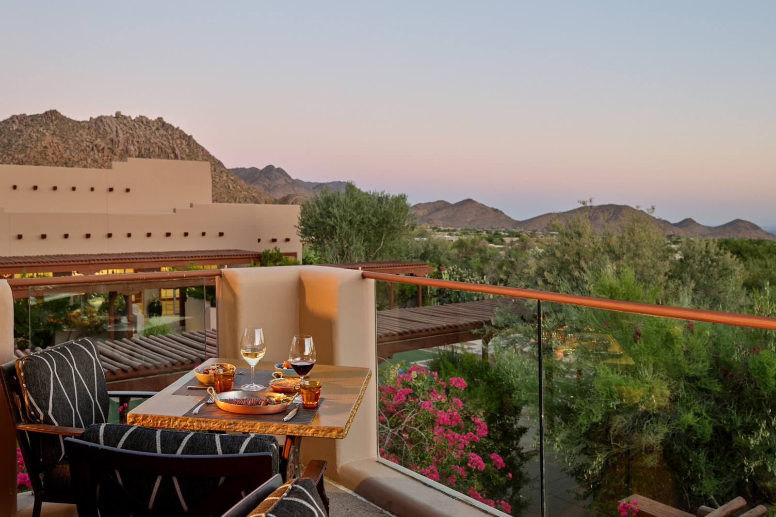 Resort balcony at dusk with wine glasses, pink sky, and rugged mountains beyond.