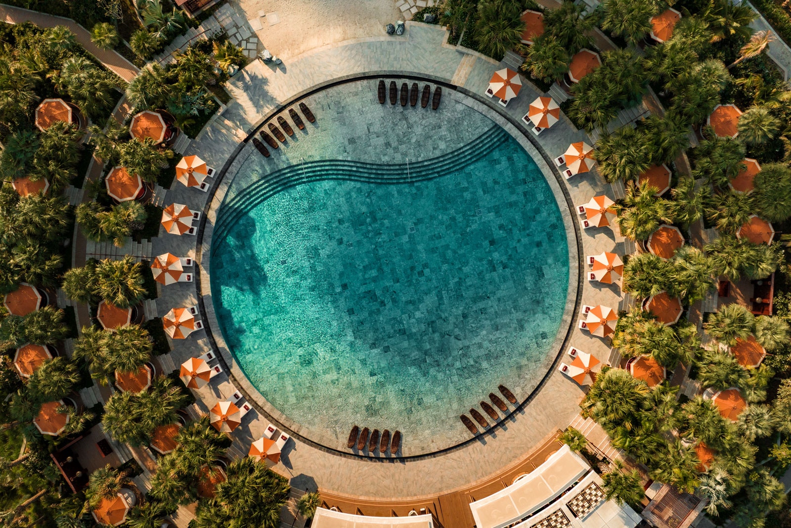 Aerial view of a round blue pool ringed by orange umbrellas amid lush green palms.