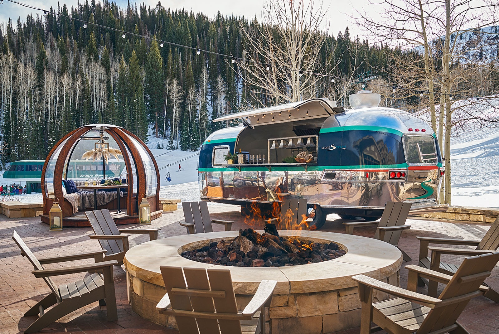 Winter patio with crackling fire pit, shiny Airstream bar, and snowy forested slopes.