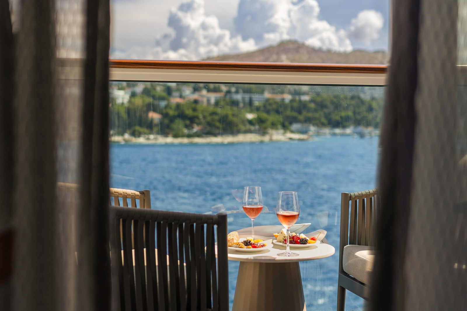 Two glasses of wine and plates of fruit on a white table, overlooking blue water and green hills from a balcony.