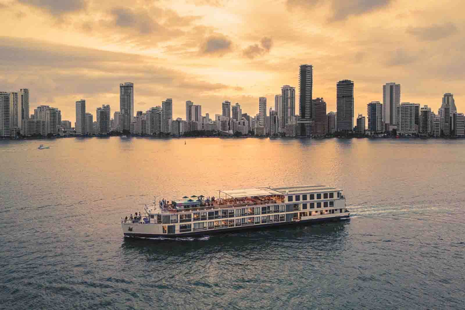 White river cruise ship sails on calm water at sunset, with a city skyline of tall gray buildings in the background.